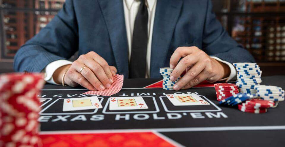 A guy in a suit sits at a gambling table, holding cards in his right hand and chips in his left.